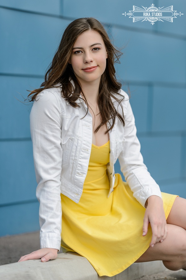 Denver senior pictures of a girl in a yellow dress, white jean jacket against a blue urban wall. 